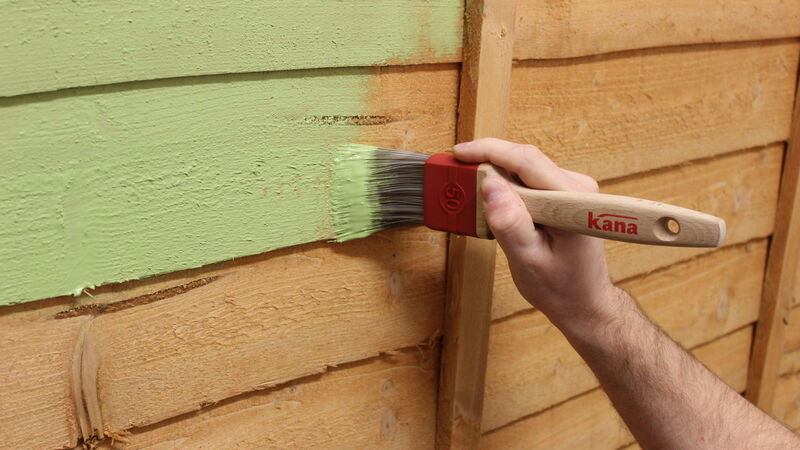 Closeup of a wooden fence being painted with a paint brush.